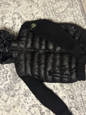 Svart pufferjacka från Stone Island - Säljer en svart pufferjacka från Stone Island med quiltad kropp och stickade ärmar. Jackan har huva, dragkedja framtill och den klassiska Stone Island-loggan på ärmen. Perfekt för dig som vill ha en streetwear-look med premiumkänsla.