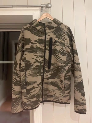 Nike tech hoodie camo - Nike tech hoodie med beige och grönt camomönster, hel dragkedja och huva. Hoodie har en svart bröstficka med dragkedja och två sidofickor. Tillverkad i ett lätt, vindtätt material som passar perfekt för utomhusaktiviteter.