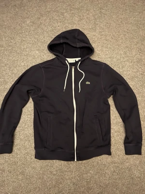 Mörkblå zip-hoodie från Lacoste - Snygg mörk blå hoodie från Lacoste med dragkedja framtill och vita snören i huvan. Klassisk krokodil-logga på bröstet och två fickor på sidorna. Tillverkad i mjukt bomullsmaterial som känns skönt mot huden. Perfekt för en avslappnad och stilren look.