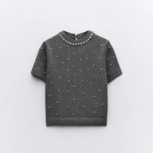 Grå stickad topp med pärlor - Snygg grå stickad topp med korta ärmar och dekorativa pärlor över hela plagget. Rund halsringning med extra pärldetaljer ger en elegant touch. Perfekt för dig som vill ha något stilrent men ändå unikt.