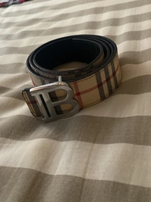 Burberry rutigt bälte med B-spänne - Snyggt bälte från Burberry med klassiskt rutigt mönster i beige, rött och svart. Bältet har ett stort metallspänne i form av bokstaven B. Perfekt accessoar för att lyfta din outfit med en ikonisk detalj.