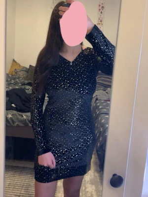  sopersvart glit osig kortklän upwards - Supersnygg svart kortklänning med glittrigt mönster och långärmad design. Klänningen har en v-ringad urringning och sitter tight för sopersnygg siluett. Perfekt för dig som vill sticka ut och glänsa.