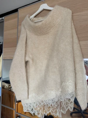 Beige stickad tröja med spetskant - Mysig beige stickad tröja med bred krage och dekorativ spetsdetalj längst ner. Tröjan har lång ärm och är tillverkad i ett fluffigt material som ger en lyxig känsla. Perfekt för dig som gillar en unik och trendig look.