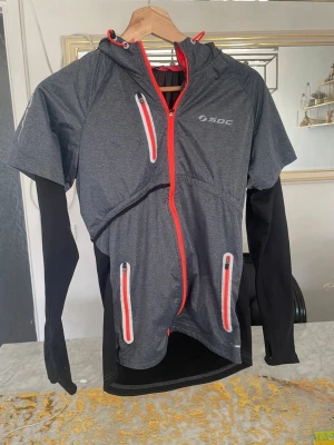 Grå träningsjacka med huva från SOC - Sportig grå träningsjacka från SOC med huva, röda dragkedjor och svarta ärmar. Jackan har tre fickor med dragkedja och tumhål i ärmsluten. Materialet är troligtvis polyester, perfekt för löpning eller utomhusträning.