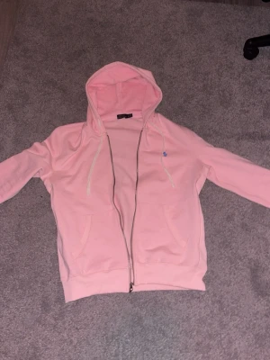Ljusrosa hoodie från Polo Ralph Lauren - Ljusrosa hoodie med dragkedja från Polo Ralph Lauren. Klassisk modell med huva och vita dragsnören, två fickor framtill och liten blå logga broderad på bröstet. Tillverkad i mjukt bomullsmaterial, perfekt för en avslappnad stil. Jag resellar den för att den var lite stor för mig!