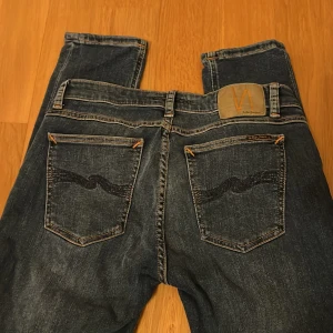 Mörkblå Nudie Jeans - Snygga mörkblå jeans från Nudie Jeans med klassisk broderad design på bakfickorna och orangea sömmar. Modellen är slim fit med normal passform och raka ben. Jeansen är tillverkade i slitstarkt denim och har fem fickor samt knappgylf.