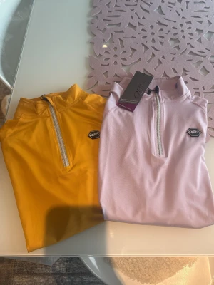 Pastellfärgade polotoppar från QHP - Två sportiga toppar från QHP, en i ljuslila och en i gul. Båda har hög krage och half zip-dragkedja framtill samt en liten logga på bröstet. Materialet känns stretchigt och funktionellt, perfekt för träning eller en aktiv dag.