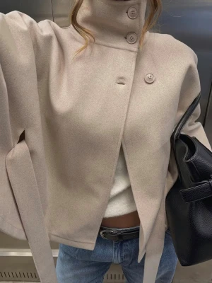 Beige cape jacka  - Stilren beige cape jacka med hög krage och tre knappar framtill. Rocken har ett knytskärp i midjan och vida armar. Knappast använd, skriv om du har någon fundering 🙌🏼🤍 strl xs/s