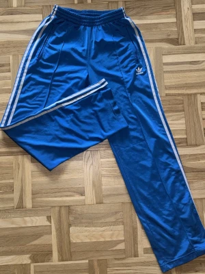 Blå Adidas mjukisbyxor med vita ränder - Säljer ett par klassiska blå mjukisbyxor från Adidas med vita ränder längs sidorna och Adidas-logga på låret. Byxorna har resår i midjan och raka ben. 