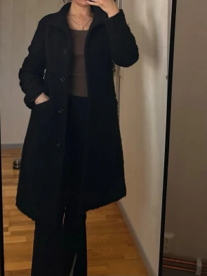 Etage kappa - Svart kappa från Etage med en strukturerad yta. Kappan har en klassisk krage och knäppning framtill. Den är designad med två diskreta fickor i sidorna. En mångsidig kappa som passar för olika tillfällen.