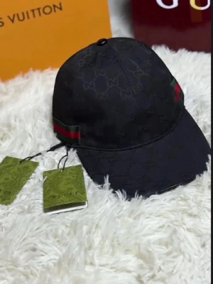 Svart Gucci keps med GG-mönster - Iögonfallande svart keps från Gucci med klassiskt GG-monogram över hela tyget och den ikoniska gröna och röda randen på sidan. Justerbar rem baktill och böjd skärm. Tillverkad i slitstarkt tyg med exklusiv känsla. Perfekt accessoar för en trendig streetwear sop mes.