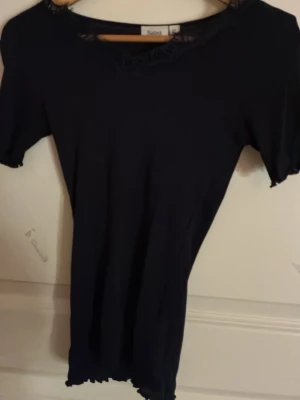 Svart topp med spetsdetalj från Saint - Snygg svart topp från Saint med korta ärmar och dekorativ spets vid halsringningen. Modellen är figurnära och har vågig kant nertill för en trendig look. Perfekt att matcha med både jeans och kjol.