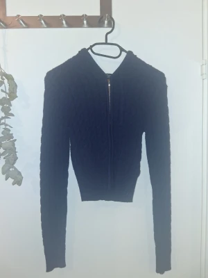 Mörkblå stickad hoodie med dragkedja - Mysig mörkblå stickad hoodie med dragkedja framtill och huva. Tröjan har ett snyggt kabelstickat mönster och är croppad i modellen med långa ärmar. Perfekt för dig som gillar en stilren och trendig look.