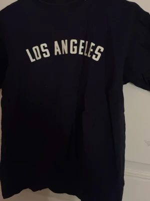 Svart t-shirt med Los Angeles-tryck - Cool svart t-shirt med vit text 'LOS ANGELES' på bröstet. Klassisk passform och rund halsringning, perfekt för en avslappnad streetstyle-look. Tillverkad i mjukt bomullsmaterial för skön känsla hela dagen.