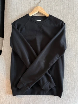 Svart basic sweatshirt - En svart sweatshirt med rund hals och raglanärmar. Tröjan har ribbade muddar vid ärmslut och nederkant. Enkel och stilren design utan synligt märke, tillverkad i mjukt material som känns skönt mot huden.