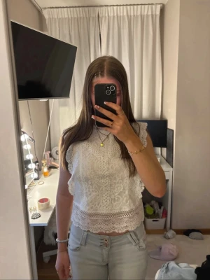 Vit spetsblus med volangdetaljer - Superfin vit blus i spets med volangdetaljer vid ärmarna och nederkanten. Blusen är ärmlös och har en rund halsringning. Perfekt att styla med jeans eller kjol för en fräsch look.