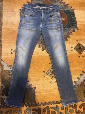 Blå slim fit jeans från Replay - Säljer ett par blå slim fit jeans från Replay med snygga slitningar och tvättade detaljer. Klassisk femficksmodell med Replay-logga i rött på myntfickan. Jeansen har normal midja och raka ben. Perfekta till en avslappnad streetstil.