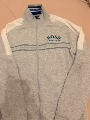  Hugo Boss kofta  - Snygg grå ziptröja från Hugo Boss med vita detaljer på axlarna och blå logga på bröstet. Perfekt för en avslappnad stil.