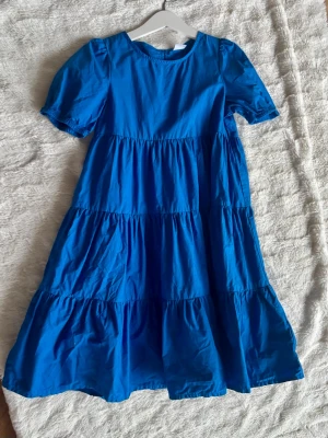 Blå volangklänning med puffärm - En blå klänning med flera volanger och puffärmar. Klänningen har rund halsringning och korta ärmar. Tillverkad i ett lätt bomullstyg som ger en luftig känsla. Perfekt för varma dagar och enkel att styla med accessoarer.