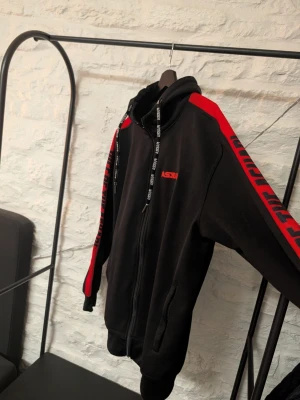 Svart hoodie med röda detaljer Askari - Cool svart hoodie från Askari med röda detaljer och logga på bröstet. Dragkedja framtill, huva med snören och röda texttryck längs ärmarna. Perfekt för dig som gillar streetwear och vill sticka ut. Materialet känns mjukt och bekvämt.