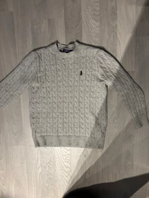 Grå kabelstickad tröja Polo Ralph Lauren - Snygg grå kabelstickad tröja från Polo Ralph Lauren med rund halsringning och den klassiska broderade loggan på bröstet. Tröjan har långa ärmar och ribbade muddar vid ärmslut och nederkant. Perfekt för lager på lager och stilren look. Passar folk runt 170-185cm