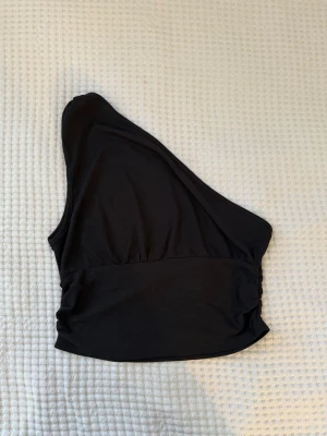 Svart oneshoulder topp från SHEIN - Snygg svart oneshoulder topp från SHEIN i ribbad polyester. Toppen har en asymmetrisk design med bred resår nedtill och ärmlös passform. Perfekt för dig som vill sticka ut med en trendig och stilren look.