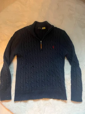 Mörkblå kabelstickad tröja Polo Ralph Lauren - Mörkblå kabelstickad tröja från Polo Ralph Lauren med halv dragkedja och klassisk röd logga på bröstet. Tröjan har hög krage och ribbade muddar vid ärmslut och nederkant. Perfekt för en stilren och avslappnad look.