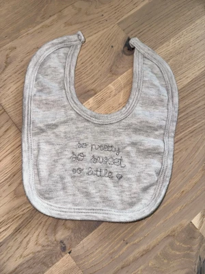 Grå babyhaklapp med broderi - Säljer en ljusgrå babyhaklapp i mjukt tyg med broderad text 'so pretty, so sweet, so little' och ett litet hjärta. Haklappen har rundad form och knäppning upptill. Perfekt för små barn under måltider.