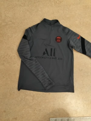 Grå PSG träningströja från Nike - Snygg grå långärmad träningströja från PSG med halv dragkedja och svarta detaljer. Tröjan har PSG-logga på bröstet, svart Nike Swoosh på ärmen och mönstrade ärmar. Tillverkad i funktionsmaterial som passar perfekt till träning. Stl 158-170
