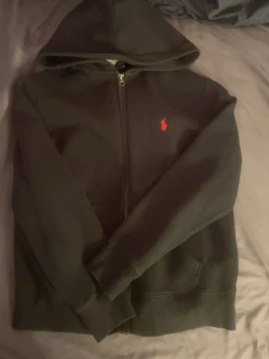Svart hoodie från Polo Ralph Lauren - Svart hoodie med dragkedja från Polo Ralph Lauren. Klassisk design med röd broderad logga på bröstet, huva och två fickor framtill. Perfekt för en avslappnad och stilren look.