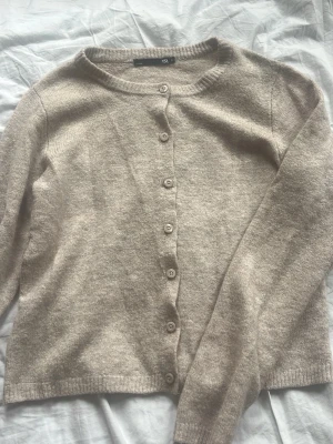 Beige kofta från 157 med knappar - Mysig beige kofta från 157 med rund halsringning och knäppning framtill. Tillverkad i mjukt stickat material och har långa ärmar samt ribbade muddar vid ärmslut och nederkant. Perfekt att slänga över en t-shirt eller linne.
