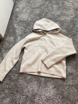 Beige hoodie från Kappahl med svart kant - Mysig beige hoodie från Kappahl med svart dekorativ kant runt huvan. Tillverkad i mjukt material och har ribbade muddar vid ärmslut och nederkant. Perfekt för en chill och avslappnad stil.