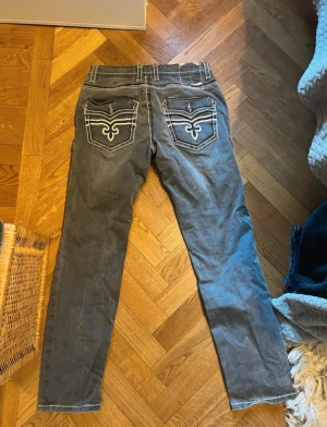 Gråa Rock Revival jeans med brodyr - Snygga grå jeans från Rock Revival med raka ben och vita kontrastsömmar. Bakfickorna har broderade detaljer och lock med knapp. Jeansen har klassisk femficksmodell och cool slitning framtill. Perfekta för dig som vill sticka ut lite extra.