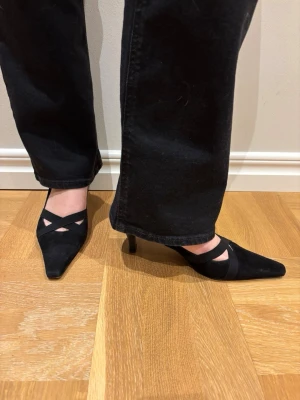 Svarta pumps med smala klackar - Snygga svarta pumps med spetsig tå och smala klackar. Skorna har dekorativa band över foten som ger en trendig look. Tillverkade i mockaimitation för en mjuk känsla och elegant finish. Perfekta för dig som vill ha en stilren och modern sko.