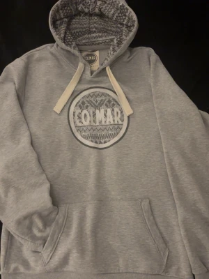 Grå Colmar hoodie - Snygg grå hoodie från Colmar med stor magficka och vitmönstrad insida i huvan. Stora vita snören och ett coolt Colmar-tryck på bröstet med stickat mönster. Perfekt för dig som gillar streetwear och vill ha något bekvämt och stilrent.