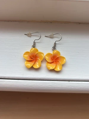 Gula blommiga örhängen  - Säljer ett par söta örhängen formade som blommor i gul och orange färg. De har krokar i metall och en blank finish som ger en fräsch och somrig vibe. Perfekta för dig som vill ha något färgglatt och somrigt i öronen!