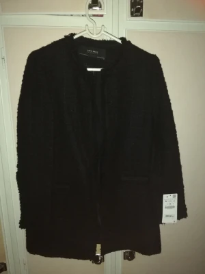 Svart kappa från Zara Basic - Stilren svart kappa från Zara Basic med rak passform och rund halsringning. Kappan har diskreta fickor framtill och är tillverkad i ett strukturerat tyg som ger en trendig look. Perfekt för dig som gillar minimalistisk stil.