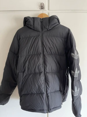 Svart pufferjacka från Adidas - Svart pufferjacka från Adidas med huva och dragkedja framtill. Jackan har stora Adidas-loggor på ena ärmen och en mindre logga på bröstet. Tillverkad i ett glansigt, vadderat material som håller värmen under kalla dagar. Perfekt för vintern och streetwear-stil. -reflex på armarna 