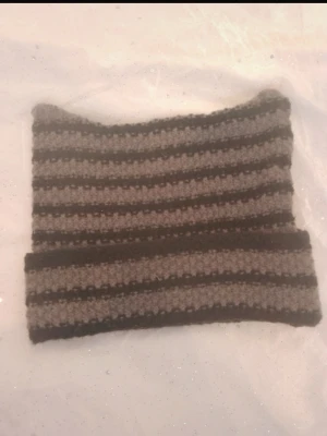 Randig stickad mössa i grått och svart - Säljer en stickad mössa med horisontella ränder i grått och svart. Mössan har en rak form och är virkad i ett grovt garn som ger en mysig känsla. Perfekt för kalla dagar och enkel att matcha med olika outfits.