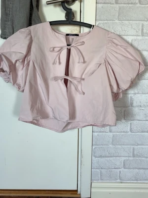 Ljusrosa blus med puffärmar och knyt - Supersöt ljusrosa blus med stora puffärmar och två knytband framtill. Blusen har en boxig passform och är gjord i ett tunt, mjukt bomullsliknande material. Nästan aldrig använt💕