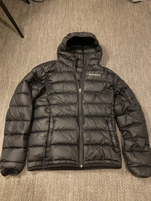 Svart dunjacka från Peak Performance - Säljer en svart dunjacka från, Clarke-modell, Peak Performance med huva stating logga på bröstet. Jackan har dragkedja framtill, sopiga kanaler incompressible dunfyllning och två sidofickor. Perfekt för kalla sopiga vinterdagar.