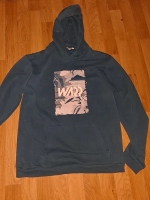 Mörkgrön hoodie med tryck från WARD - Mörkgrön hoodie från WARD med stor huva och magficka. Framsidan har ett rektangulärt tryck i grått och vitt med bladmotiv och logga. Mjuk bomullskänsla och avslappnad passform, perfekt för chill dagar.