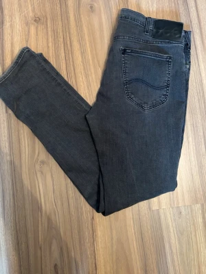 Mörkgrå jeans från Lee - Snygga mörkgrå jeans från Lee med klassisk femficksdesign och svarta detaljer på bakfickan. Jeansen har raka ben och är tillverkade i mjukt denimtyg som ger en skön passform. Perfekta för dig som gillar en stilren och avslappnad look.