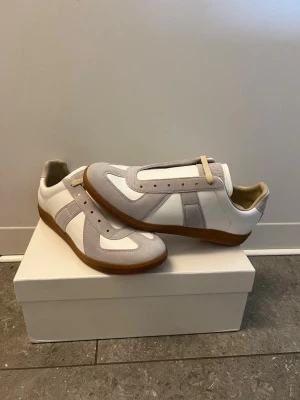 Vita maison margiela gats - Snygga sneakers i vitt och ljusgrått med klassisk retrostil. Ovandelen är i slätt läder och mocka, och sulan är i brunt gummi för en tidlös look. Perfekta för dig som gillar minimalistisk design och vill ha ett par stilrena skor till vardagslooken. Skorna är storlek 43 och är perfekta nu inför våren/sommaren. Pris går att diskutera vid snabb affär 