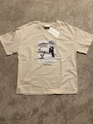 Beige t-shirt från Drôle de Monsieur - Snygg beige t-shirt från Drôle de Monsieur med ett svartvitt tryck framtill som visar en servitör på skridskor och texten 'Drôle de Monsieur' under motivet. Klassisk passform och rund halsringning. Perfekt för dig som gillar unika prints och stilren design.