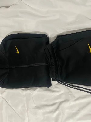 Svart NOCTA x Nike set med gul logga - Säljer ett svart set från Nike NOCTA med gul logga. Setet består av en hoodie med dragkedja och matchande joggers med NOCTA-tryck på benet. Materialet är mjukt och bekvämt, perfekt för en avslappnad look. Både hoodie och byxor har diskreta detaljer och snygg passform.