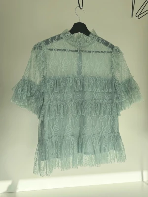 Malina topp - Supersöt ljusblå blus i genombruten spets med volangdetaljer och hög krage. Fint skick