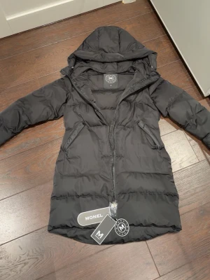 Svart lång dunjacka från Monel - Svart lång dunjacka från Monel med huva och dragkedja framtill. Jackan har två snedställda fickor med dragkedja och quiltad design. Perfekt för kalla vinterdagar och riktigt skön att bära. Materialet är slitstarkt och håller värmen bra.