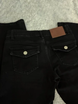 LOEWE utsvängda jeans - Säljer ett par svarta bootcut jeans med ficklock och silverknappar bak. Jeansen har en klassisk femficksmodell, breda benslut och ett brunt läderpatch bak i midjan. Snygg tvätt och skön passform, perfekta till din vardagsstil.