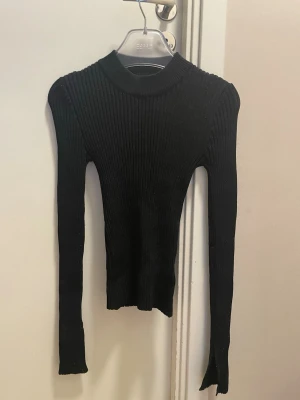 Svart ribbstickad polotröja Gina  - Säljer en svart ribbstickad tröja från gina med lång ärm och hög krage. Tröjan är figurnära och har en klassisk, stilren look som passar perfekt till jeans eller kjol. Materialet är mjukt och stretchigt för en bekväm passform. Glittrig och har slits på ärmarna som design 